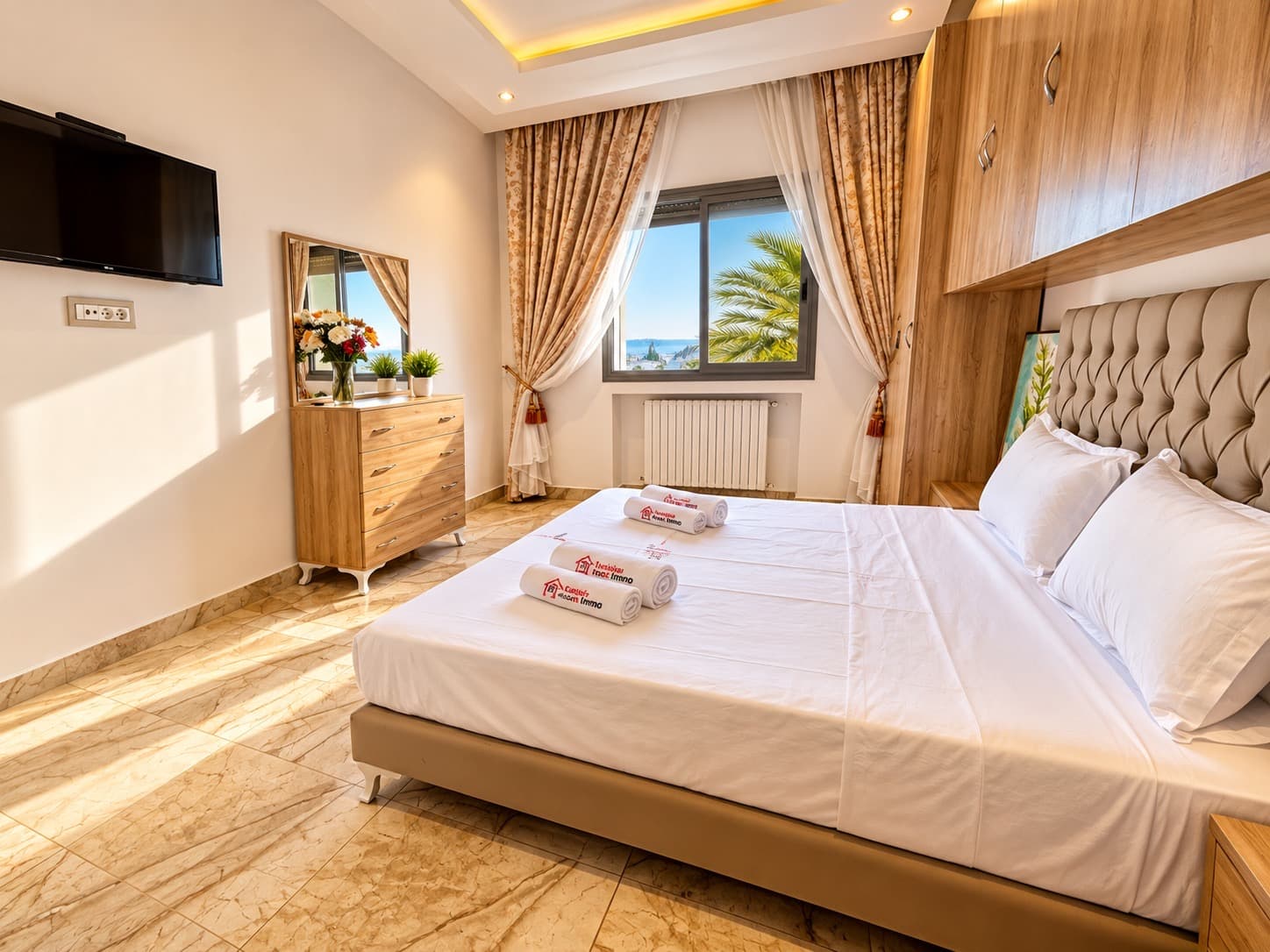 Chambre avec tête de lit capitonnée taupe, rangements en bois clair et vue sur la mer